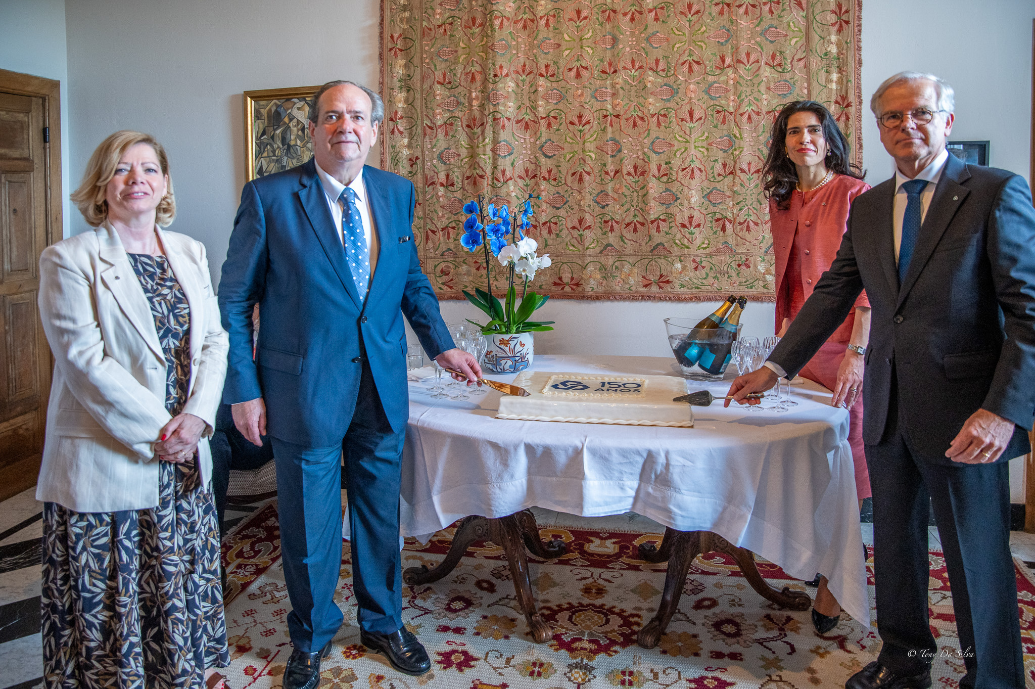 Quatro pessoas em traje formal posam junto a uma mesa decorada com flores e objetos comemorativos, numa sala elegante, durante a celebração dos 150 anos da Caixa Geral de Depósitos na Residência Oficial do Embaixador de Portugal na Bélgica.