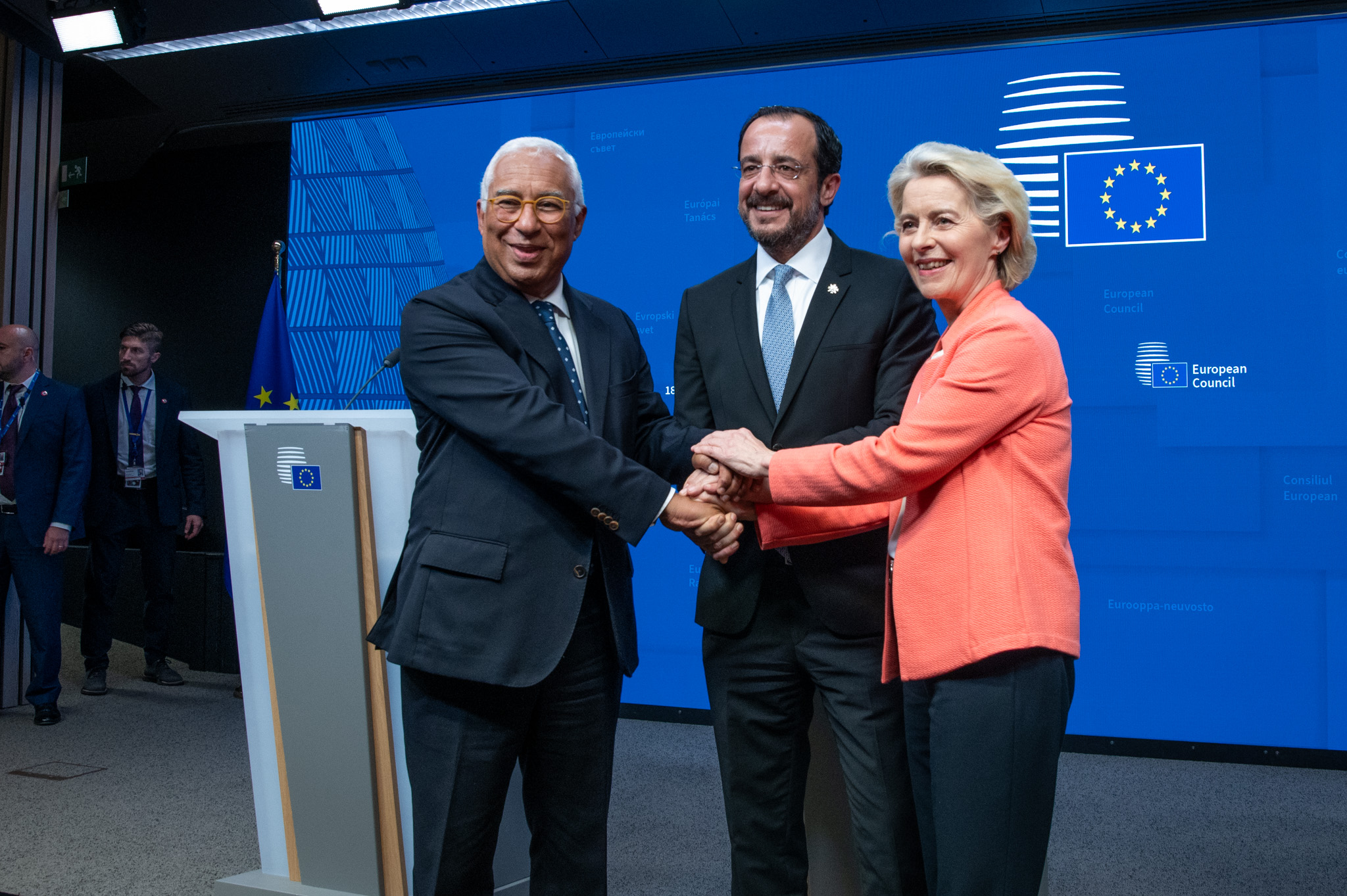 Três líderes europeus apertam as mãos em frente a um fundo azul com o logótipo da União Europeia, simbolizando unidade e cooperação após a reunião do Conselho Europeu em Bruxelas.
