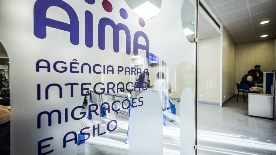 Entrada de um balcão da Agência para a Integração, Migrações e Asilo (AIMA), com o nome da agência visível no vidro e pessoas a serem atendidas no interior.