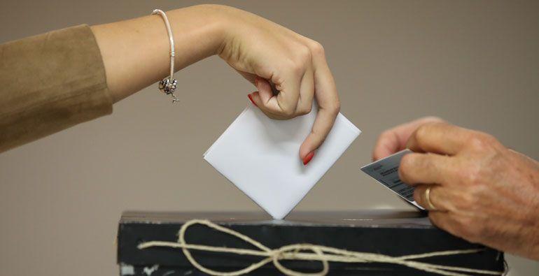 Mão de uma pessoa a colocar um boletim de voto numa urna, ilustrando o ato de votar, tema central do artigo sobre as dificuldades dos portugueses no estrangeiro em participar nas eleições.