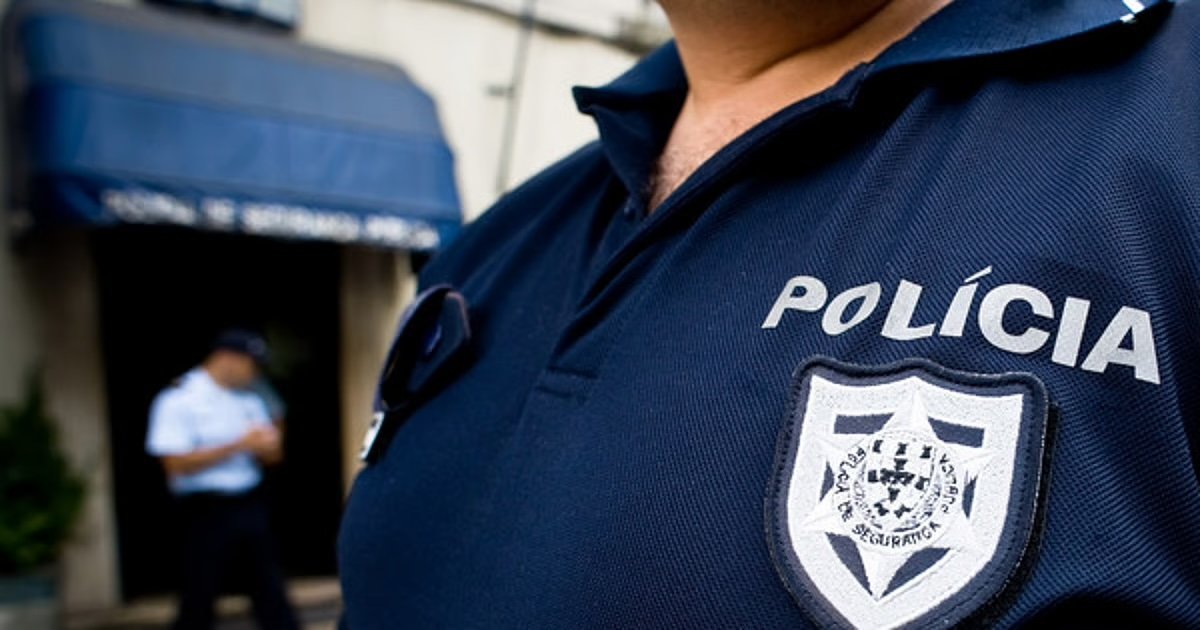 Agente da polícia com uniforme azul escuro identificado com a palavra 'Polícia' e emblema, em frente a um edifício, ilustrando ação policial.