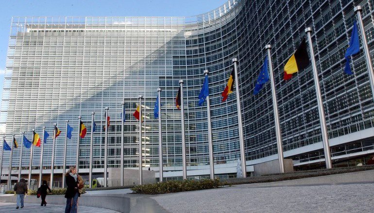 Edifício moderno da Comissão Europeia com várias bandeiras de países da União Europeia hasteadas à frente.