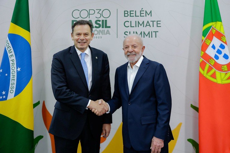 Dois líderes de fato escuro apertam as mãos em frente às bandeiras do Brasil e de Portugal, com um fundo que exibe o logótipo da Cúpula de Belém e referência à COP30.