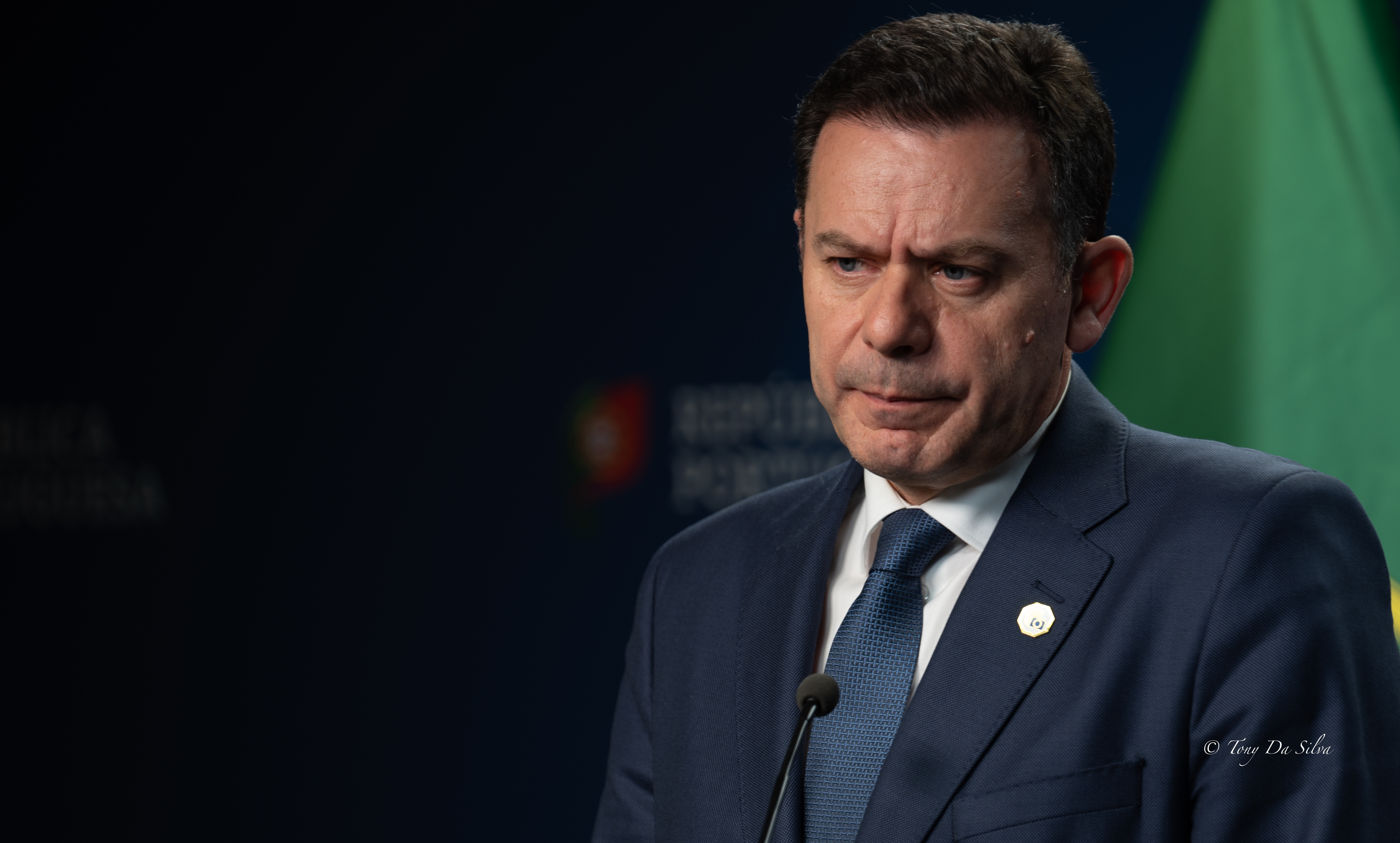 Primeiro-ministro português, Luís Montenegro, durante uma intervenção oficial, com expressão séria, ao lado de uma bandeira nacional.