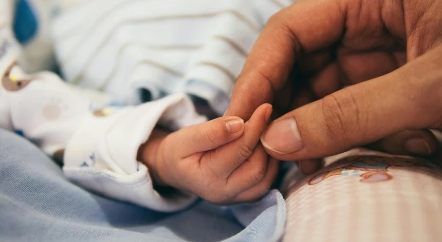 Mão de adulto a segurar suavemente a mão de um bebé, simbolizando ligação e proteção familiar.