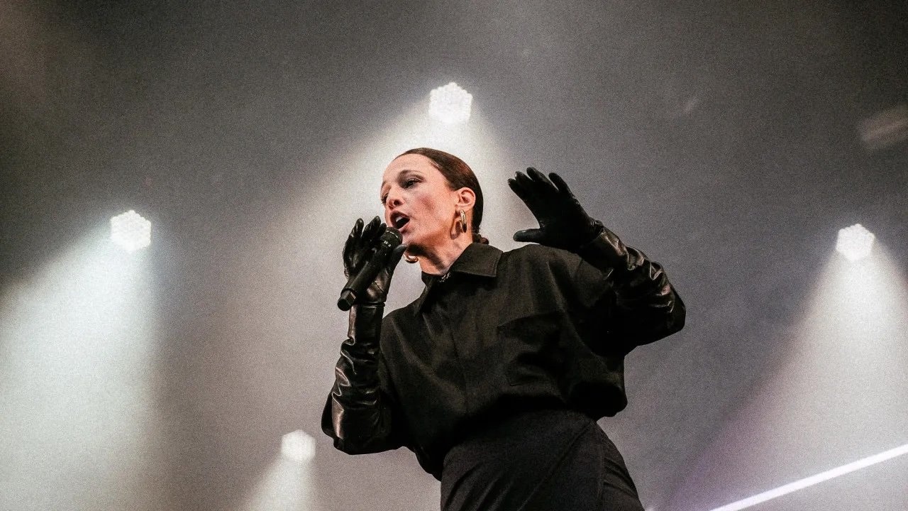 Um artista no palco, vestindo roupas pretas e luvas, cantando em um microfone com luzes brilhantes ao fundo.