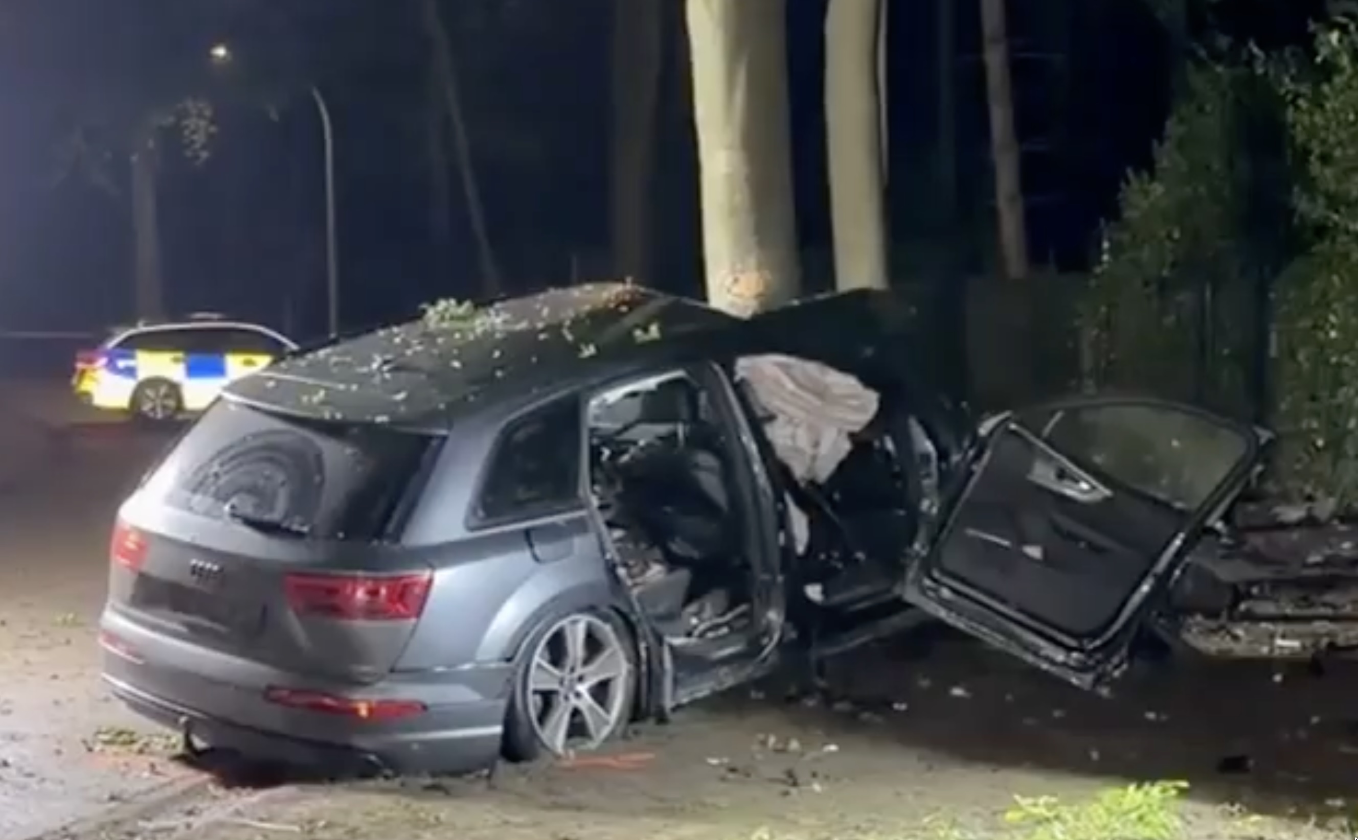Carro gravemente danificado após embater em árvores à noite, com portas abertas e presença de viatura policial ao fundo, ilustrando o acidente mortal em Brasschaat descrito no artigo.
