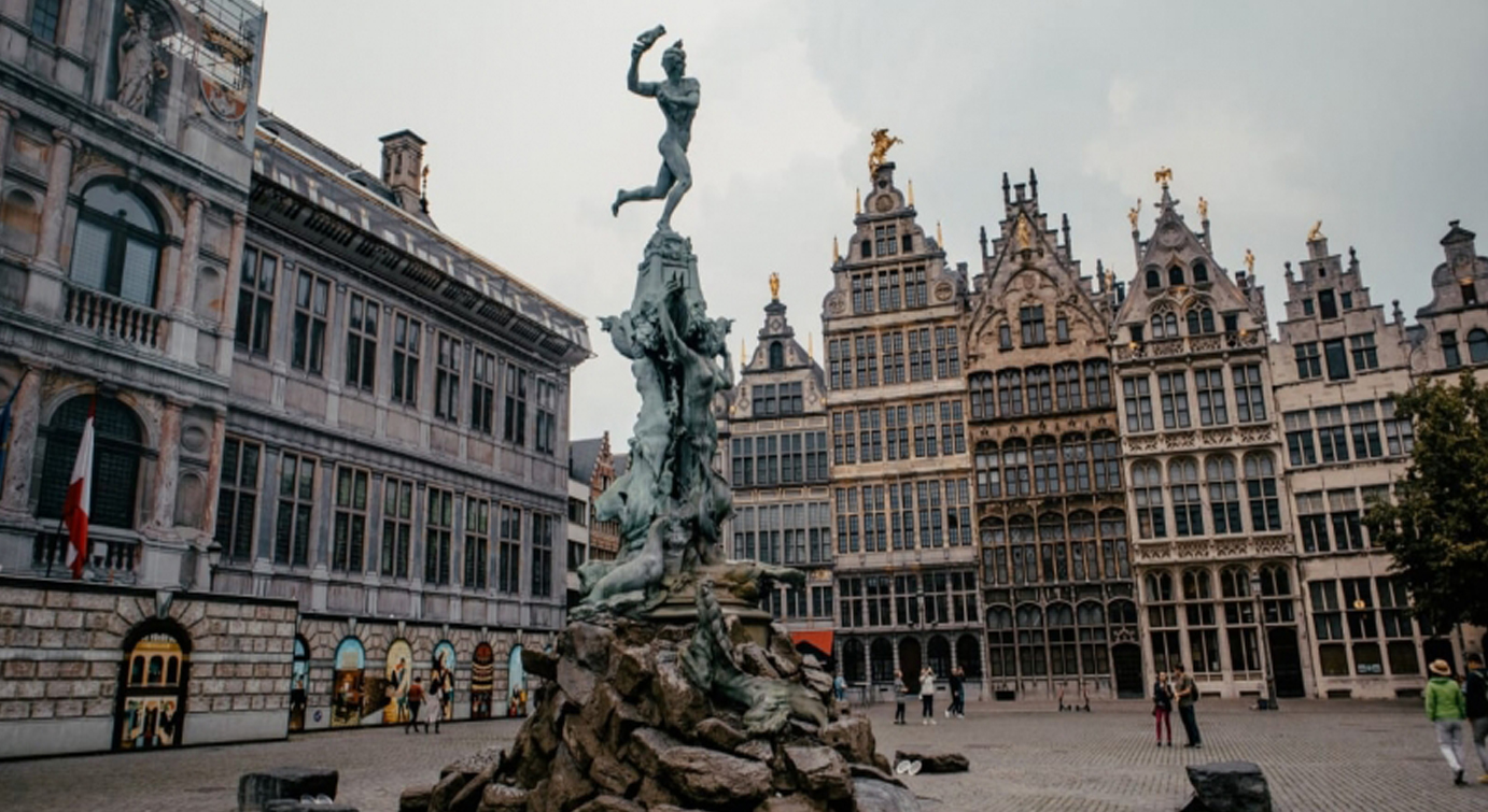 Praça central de Antuérpia com uma estátua no centro e edifícios históricos ao fundo, local onde será celebrada a festa do Dia de Portugal para a comunidade portuguesa.