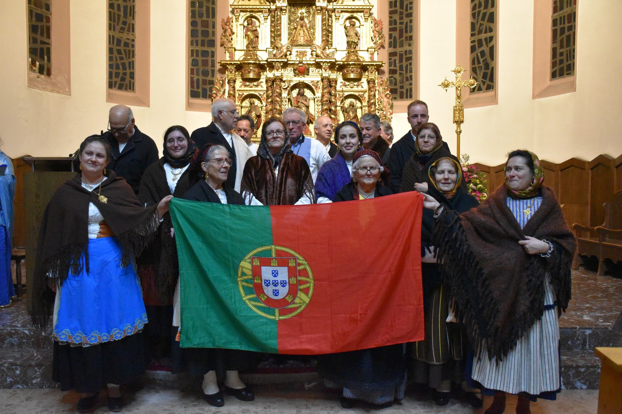 Grupo de pessoas com trajes tradicionais portugueses segura uma bandeira de Portugal no interior de uma igreja, celebrando a tradição das Janeiras.