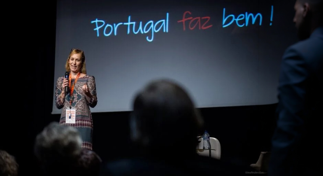 Oradora em palco durante um evento, com a frase 'Portugal faz bem' projetada no ecrã atrás, perante uma audiência.