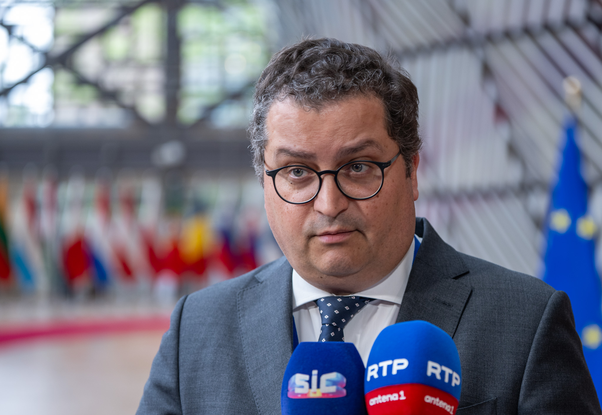 O ministro das Finanças português, de fato e gravata, fala à imprensa em Bruxelas, com microfones de canais portugueses à sua frente e bandeiras da União Europeia e outros países ao fundo.