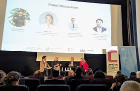 Painel de discussão com quatro pessoas sentadas em palco, moderador à esquerda, ecrã de projeção ao fundo com nomes e fotos dos oradores, público sentado a assistir. Evento celebra a língua portuguesa em Bruxelas.