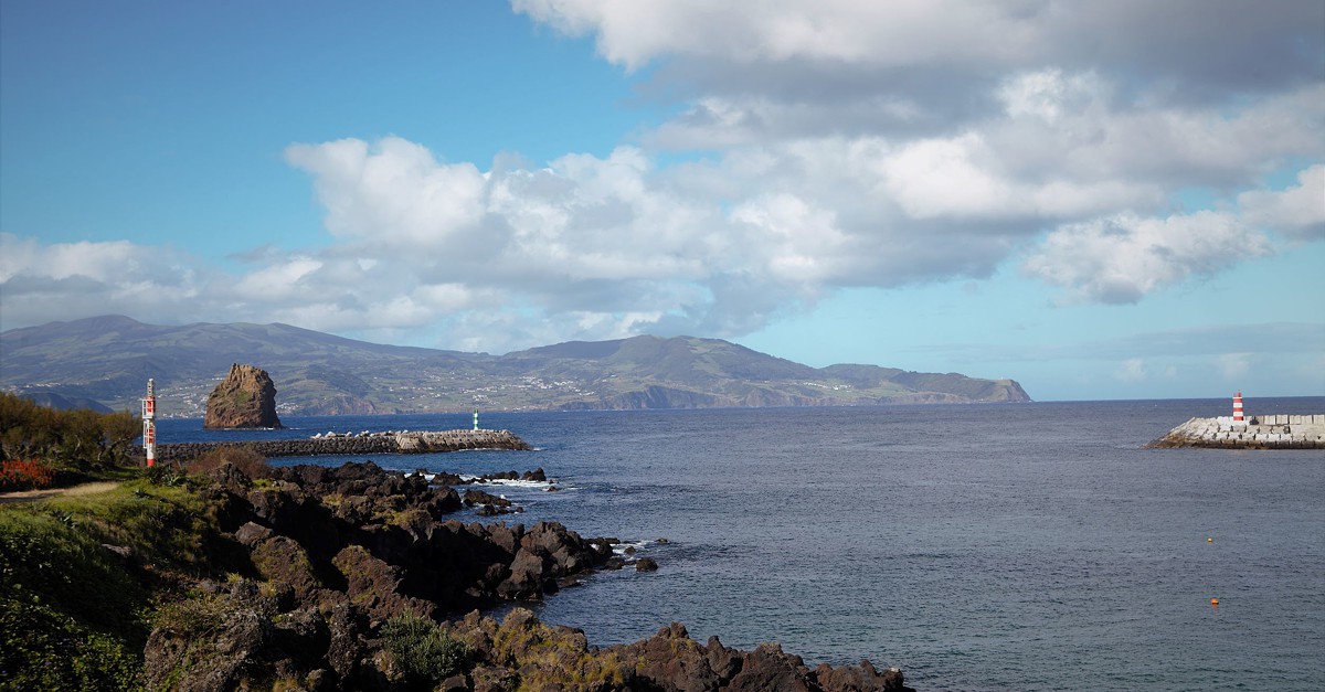 Vista costeira do canal entre o Faial e o Pico, Açores, com mar calmo, costa rochosa, duas boias de sinalização e montanhas ao fundo sob céu parcialmente nublado.