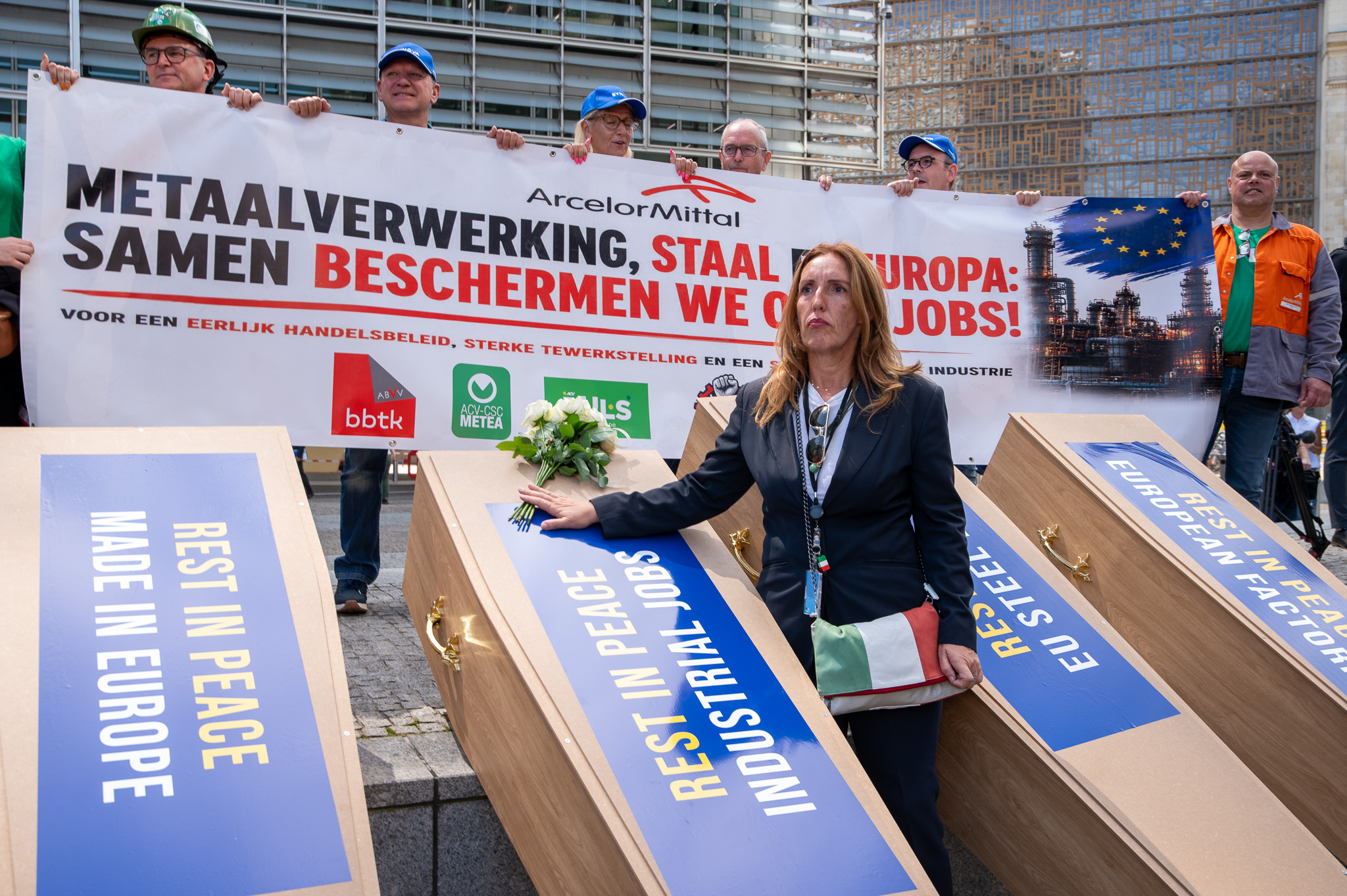 Manifestação em Bruxelas com representantes do setor metalúrgico junto a caixões simbólicos que têm mensagens como 'Rest in Peace Made in Europe', e um cartaz ao fundo a exigir proteção para a indústria e os empregos europeus.