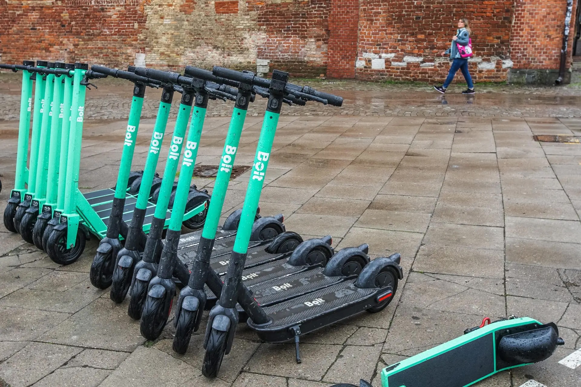 Várias trotinetes elétricas de aluguer alinhadas numa rua de Bruxelas, com uma pessoa a caminhar ao fundo junto a uma parede de tijolo.