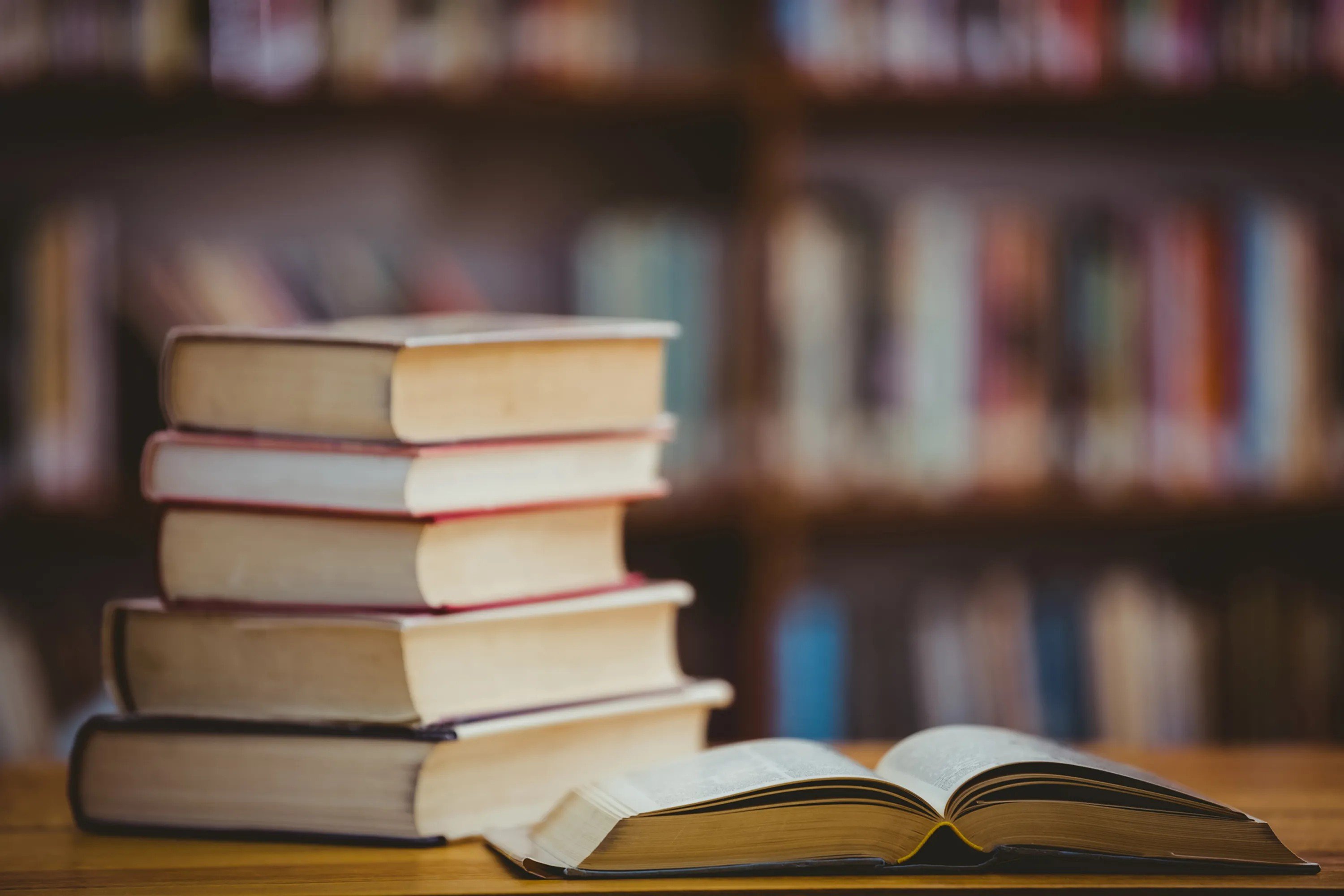 Pilha de livros e um livro aberto sobre uma mesa, com estantes desfocadas ao fundo, sugerindo um ambiente literário.