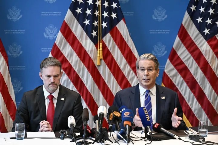 Conferência de imprensa com dois homens em fato e gravata sentados à mesa, rodeados por microfones, com bandeiras dos Estados Unidos ao fundo, representando uma comunicação oficial do governo norte-americano.