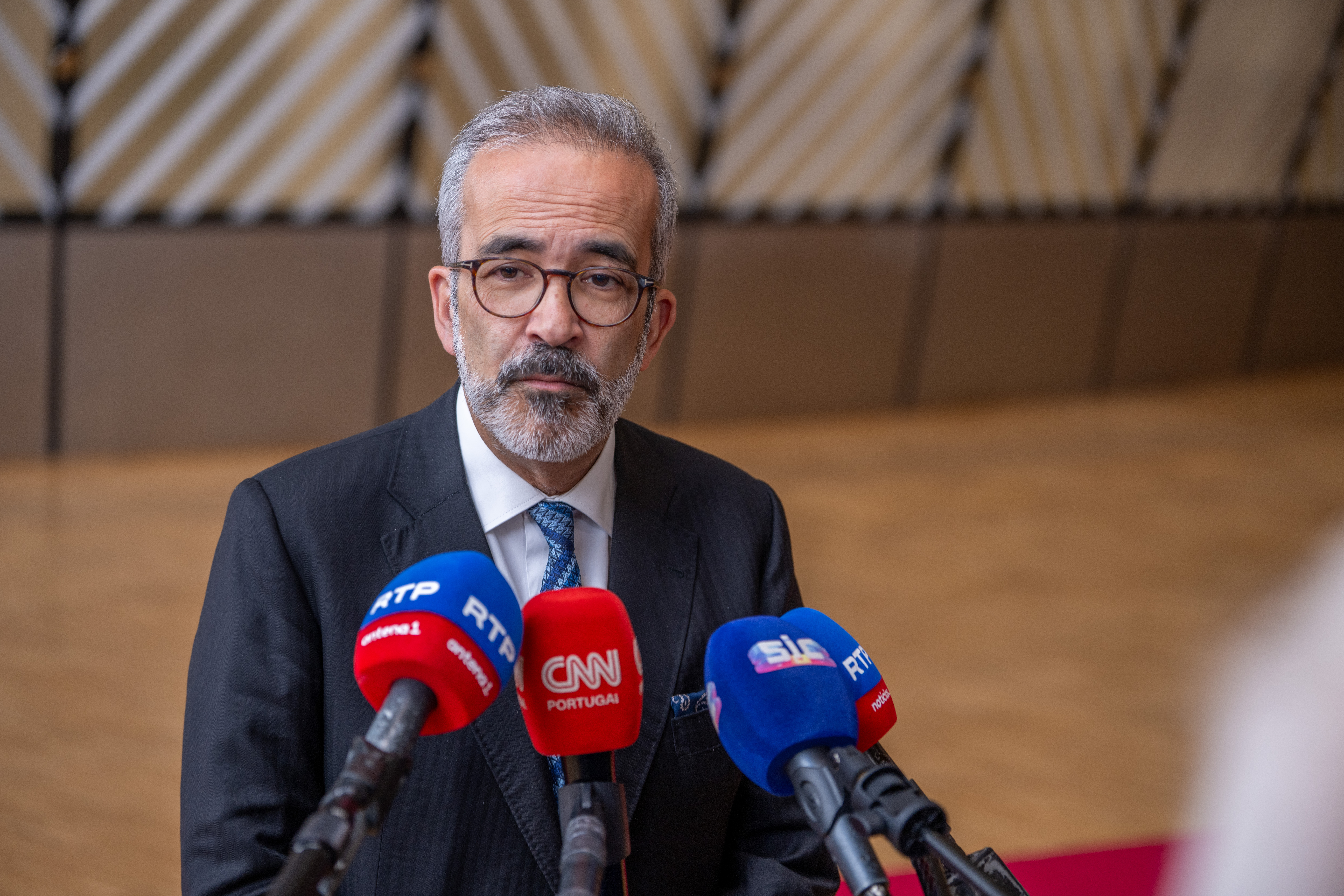 O ministro dos Negócios Estrangeiros português fala à imprensa, rodeado de microfones de vários meios de comunicação, durante uma conferência em Bruxelas sobre sanções da União Europeia a Israel.